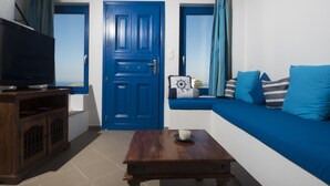 A room with a blue door, a television on a wooden stand, a wooden table with a candle, and a blue sofa with cushions. - Santorini Traditional Suites (Santorini)