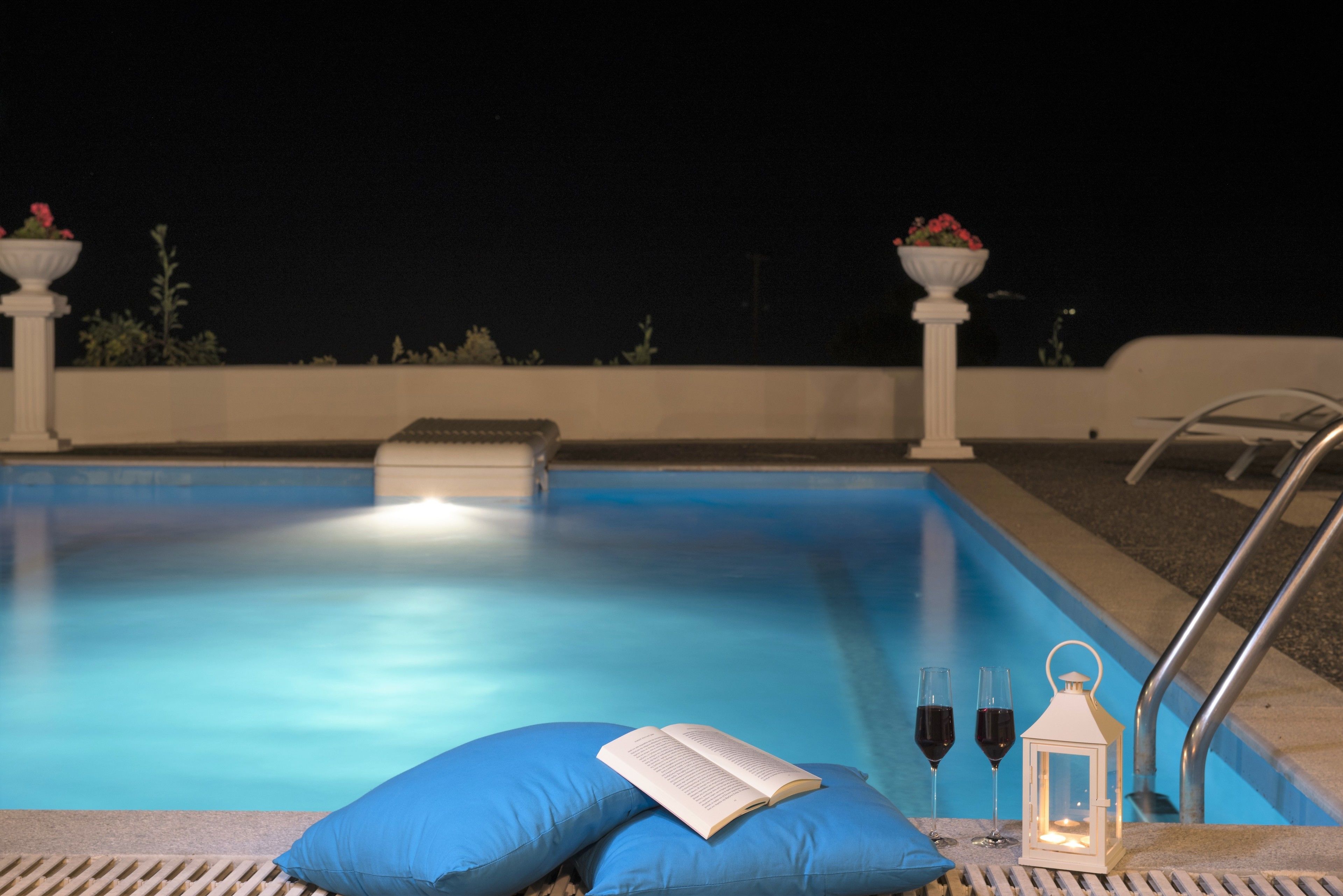 A poolside area with a blue lounge chair, a book, two wine glasses, and a lantern.