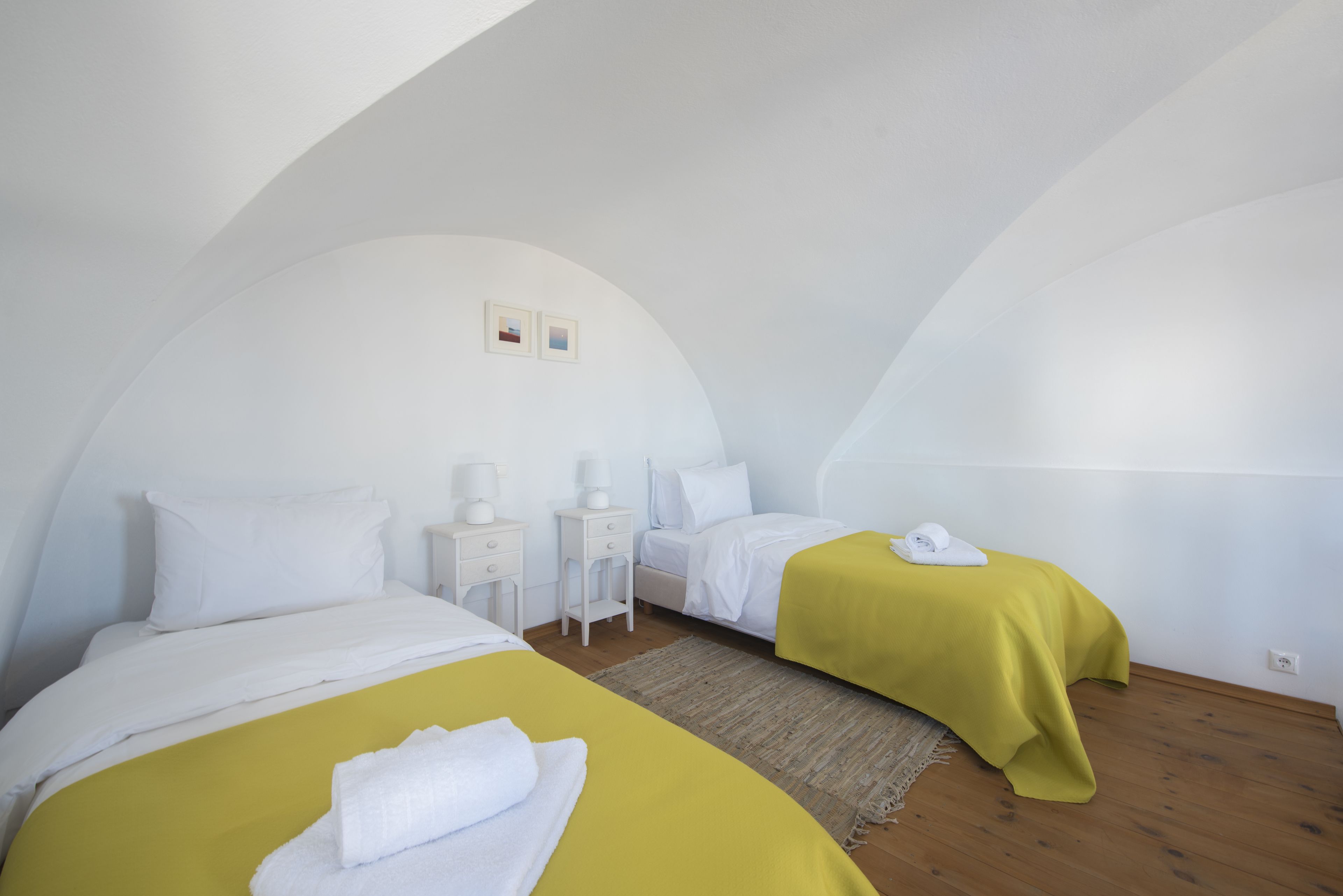 A room with two beds, each with a yellow bedspread and white pillows, a small bedside table with a lamp, and a wall-mounted picture.