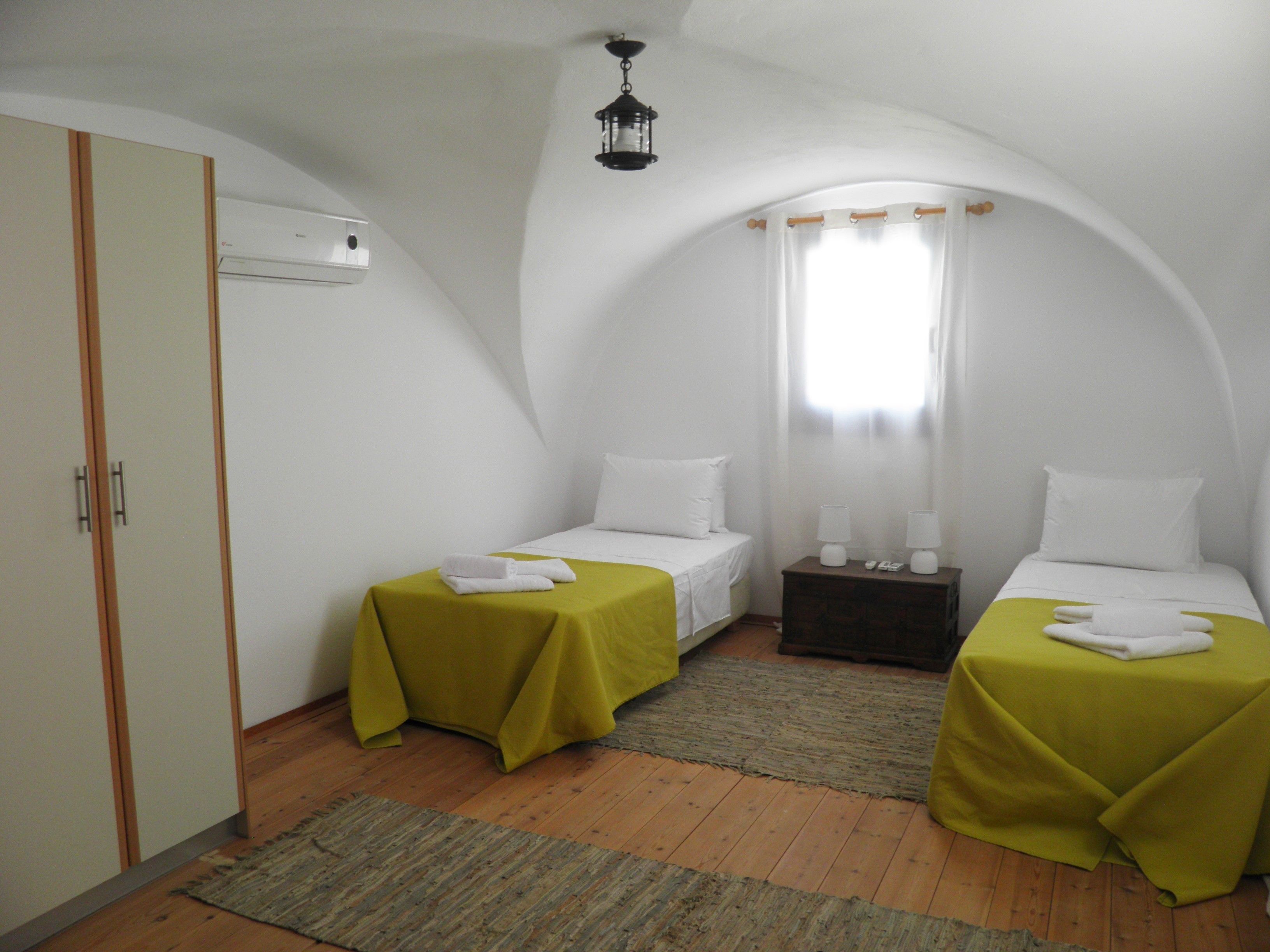 A small, white, two-bed room with green bedspreads, wooden flooring, and a single window with curtains.