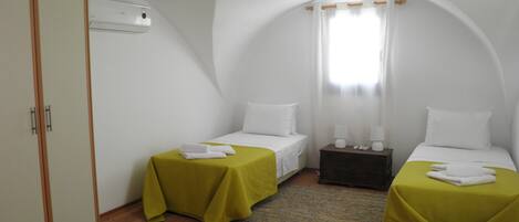 A small, white, two-bed room with green bedspreads, wooden flooring, and a single window with curtains.