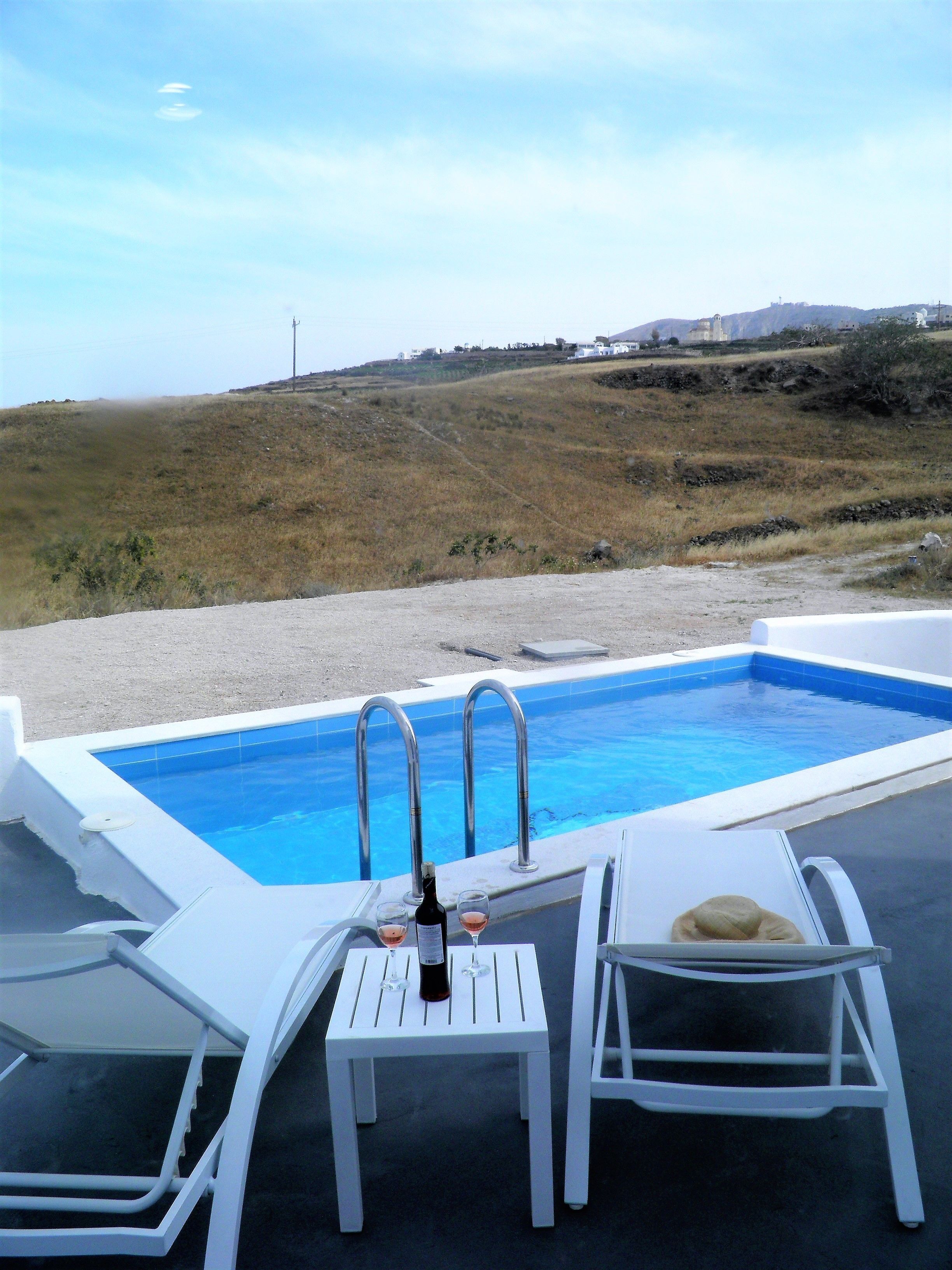 A pool area with a table and chairs, a bottle of wine, and a view of the landscape.