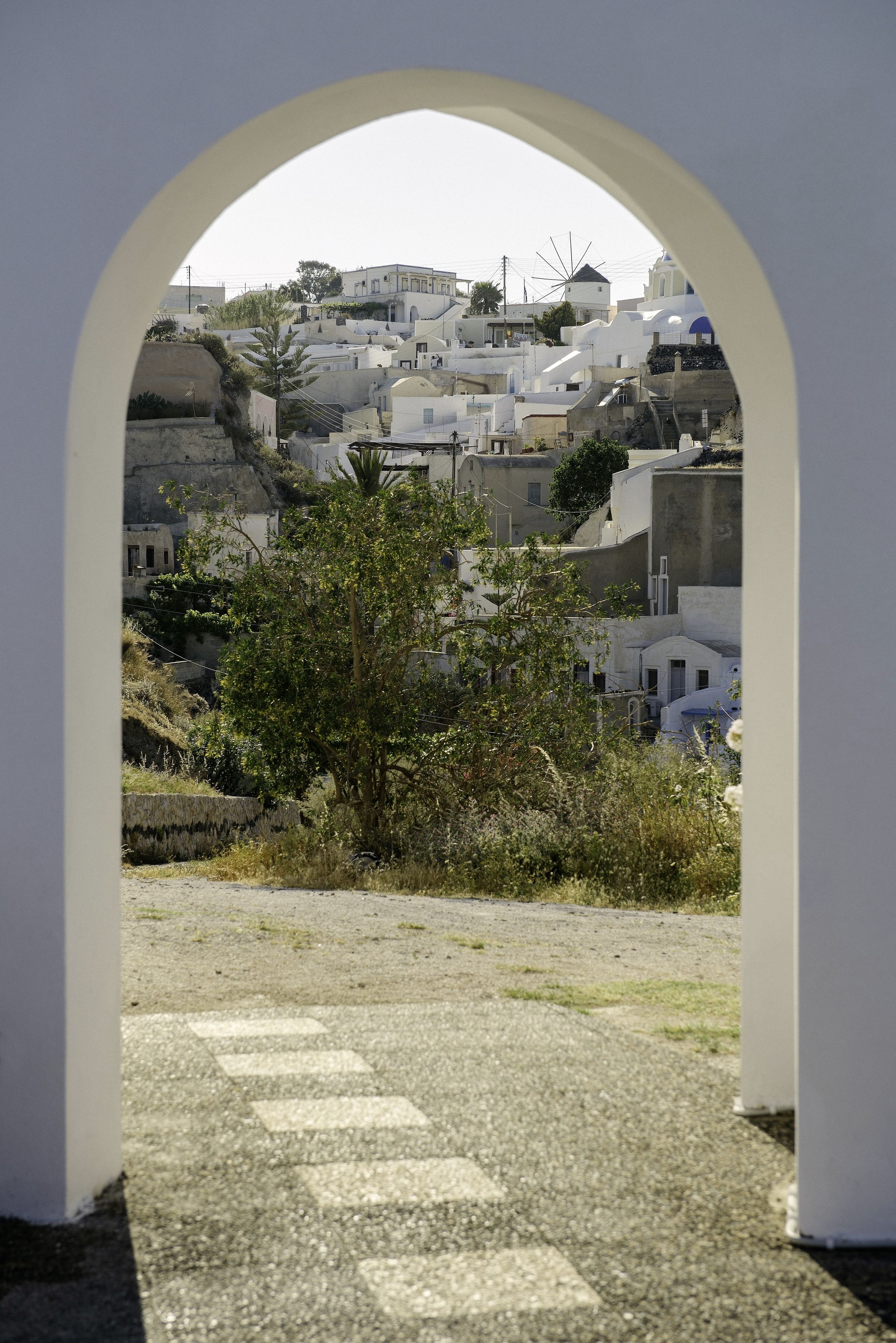 A view through an archway of a coastal town with white buildings, a tree in the foreground, and a clear sky.