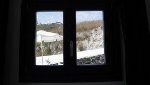 A window with a view of a white building on a hill, a stone wall, and a grassy area. - Santorini Traditional Suites (Santorini)