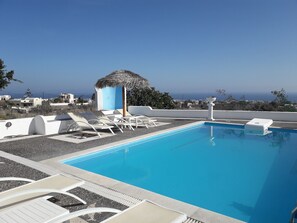 Ein Poolbereich mit Liegestühlen und einer Struktur mit Reetdach, umgeben von einem weißen Gebäude und Blick aufs Meer.