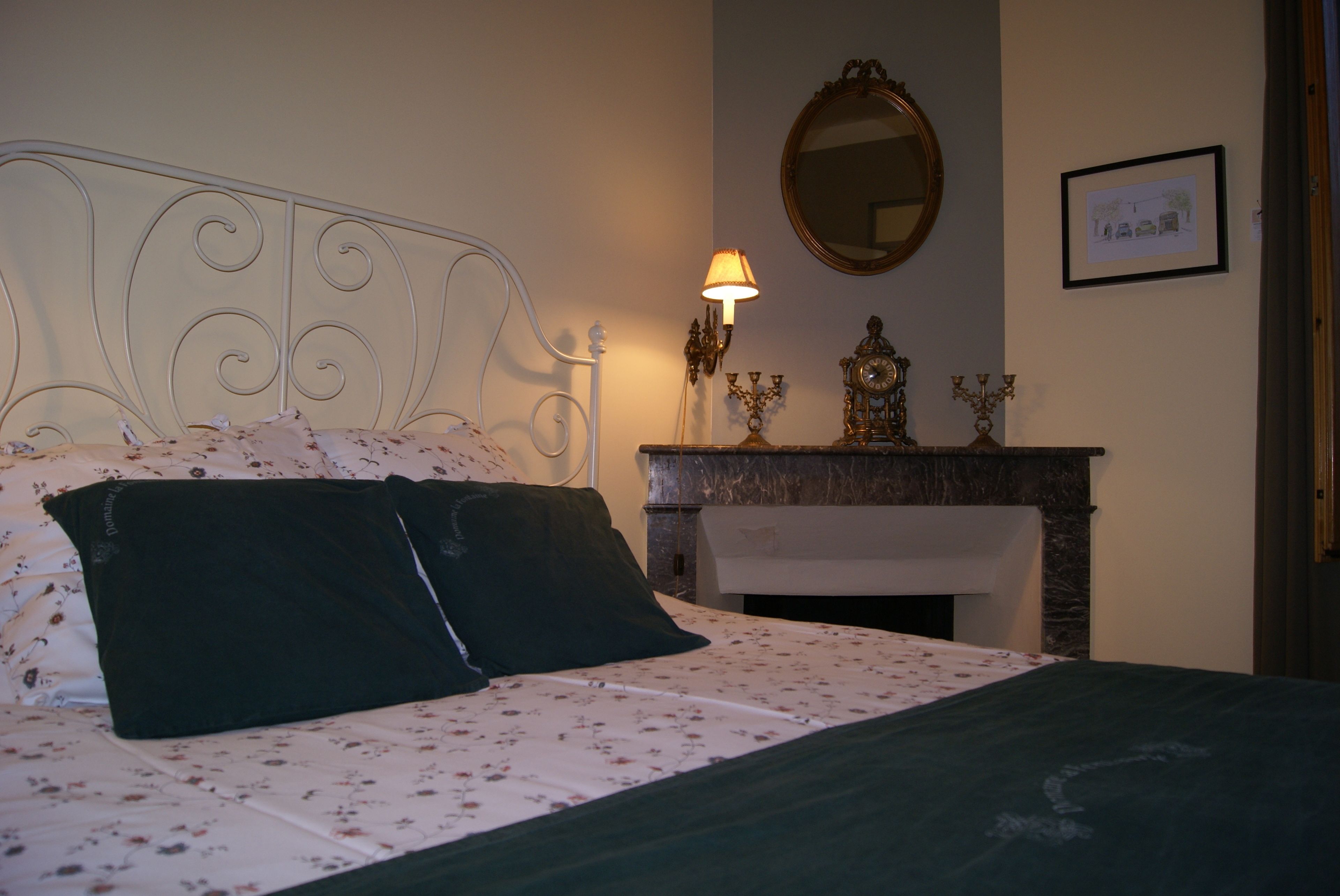 A neatly made bed with a floral bedspread and two pillows, a wall-mounted mirror, a framed picture, and a fireplace with decorative items.