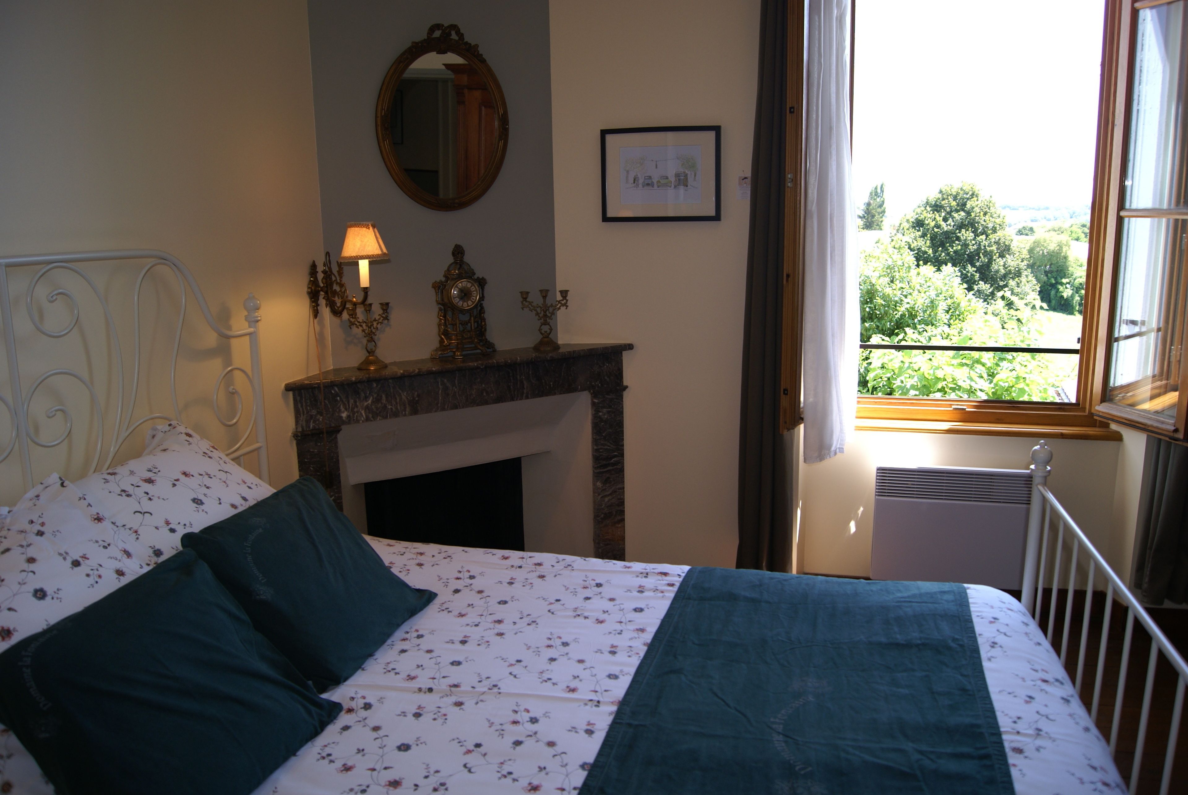 Un dormitorio con cama, chimenea, espejo y ventana con vista a vegetación.