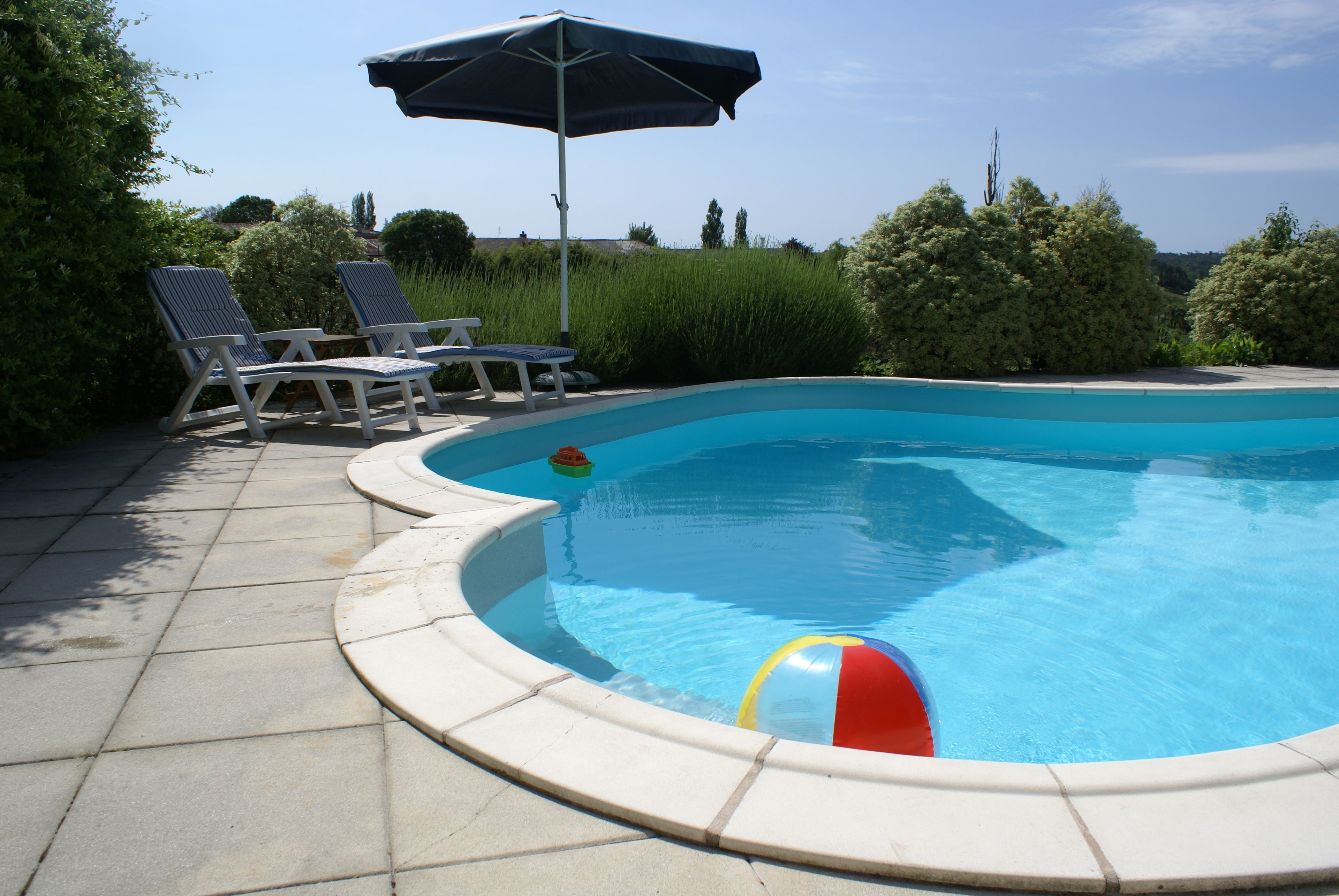 Ein Pool mit gewölbter Kante, umgeben von einer gefliesten Liegefläche. Zwei Liegestühle und ein Strandball liegen in der Nähe.