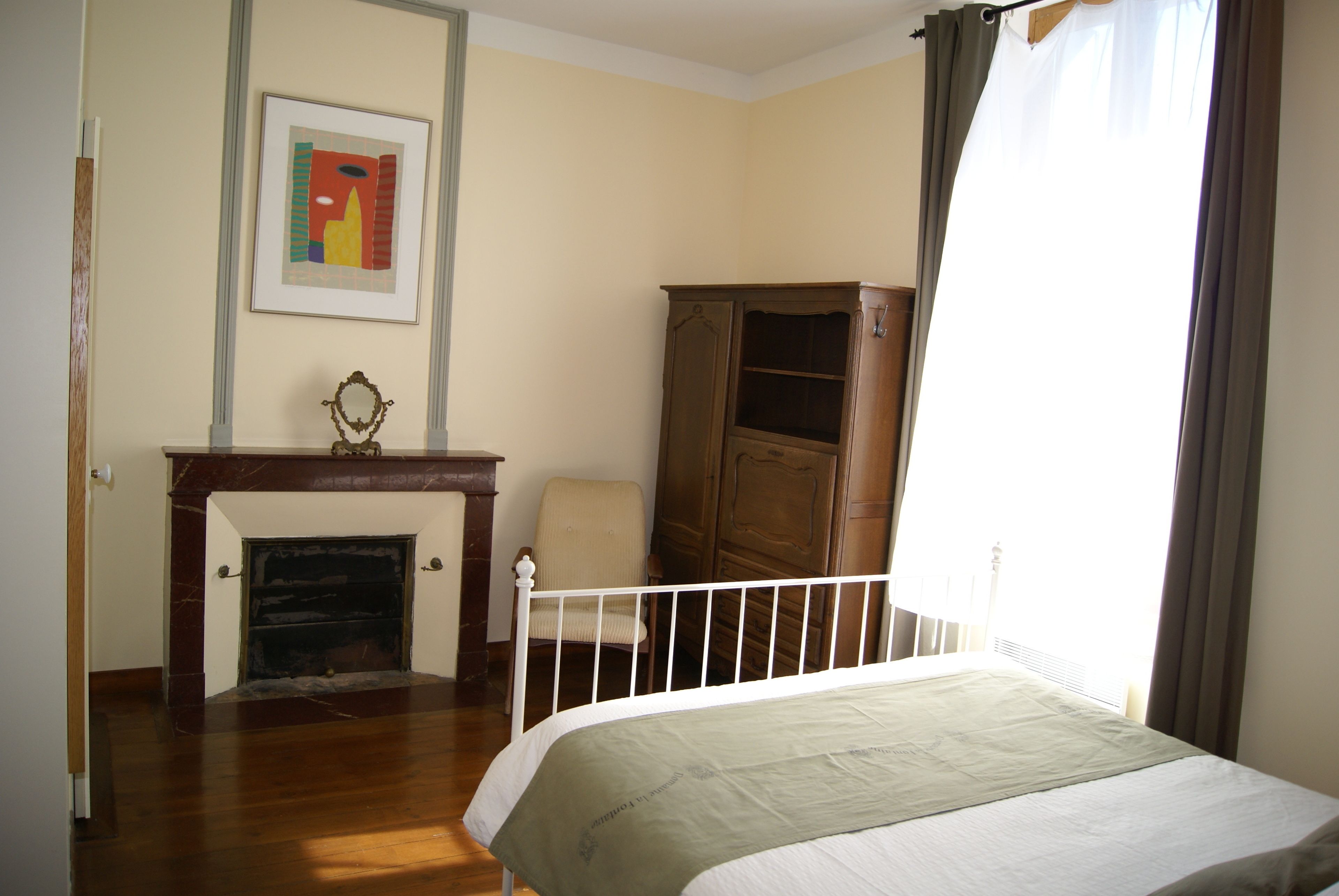 A bedroom with a bed, a wooden wardrobe, a chair, a fireplace, and a framed picture on the wall.