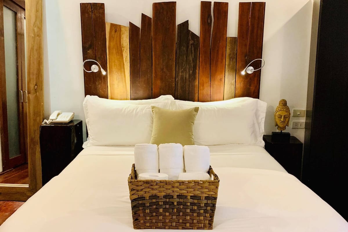 A neatly made bed with white linens, a wicker basket with towels, and a wooden headboard with vertical slats.