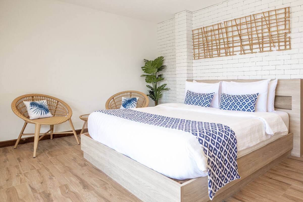A bedroom with a bed, two wicker chairs, a potted plant, and a white brick wall.