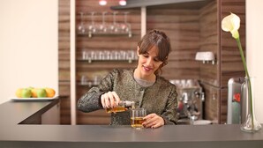 Una persona sirve una bebida en un vaso en un bar, con un fondo de madera y un jarrón con una flor.