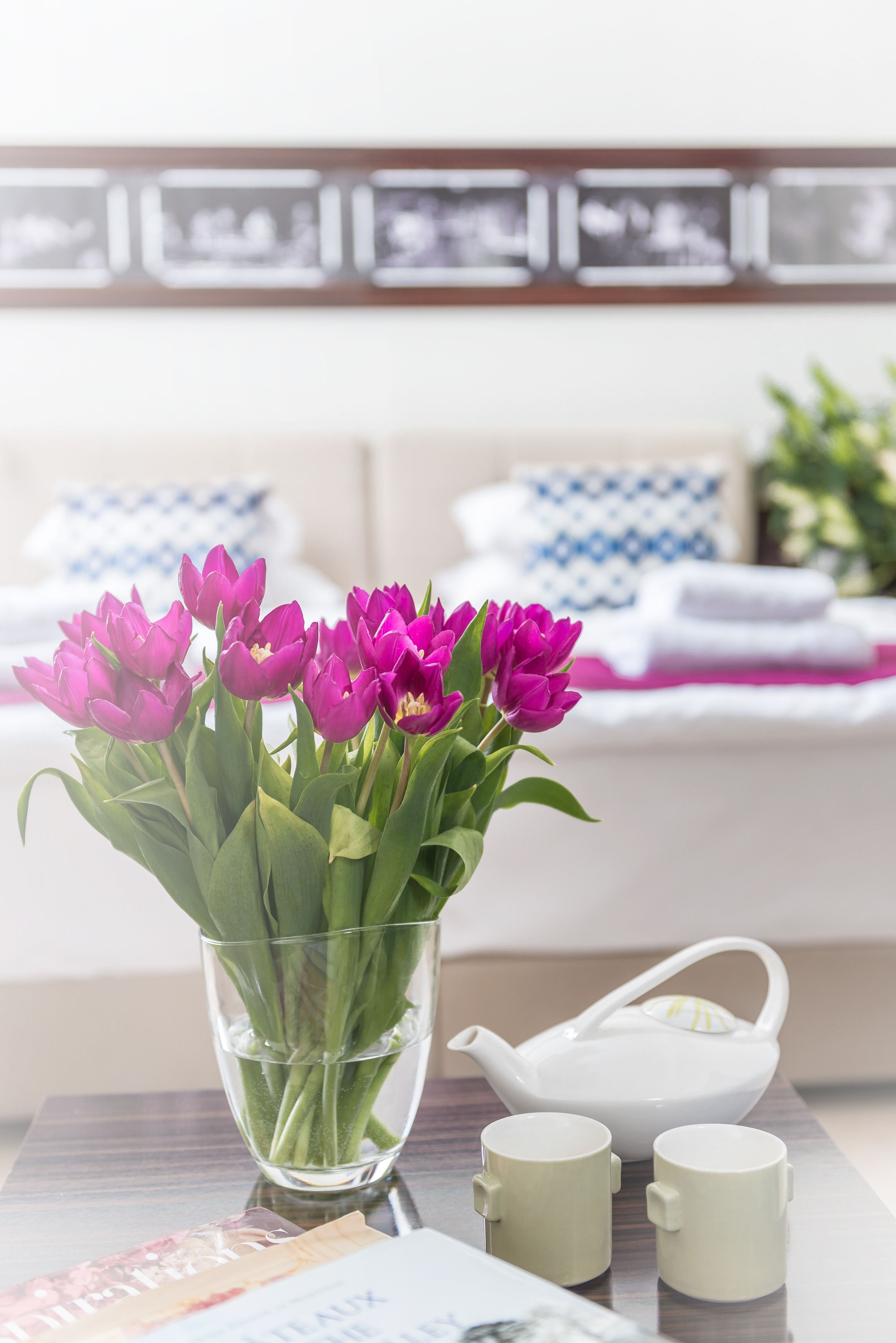 Un vaso di vetro trasparente con tulipani viola intensi su un tavolo, insieme a due tazze di ceramica abbinate e un teiera bianca.