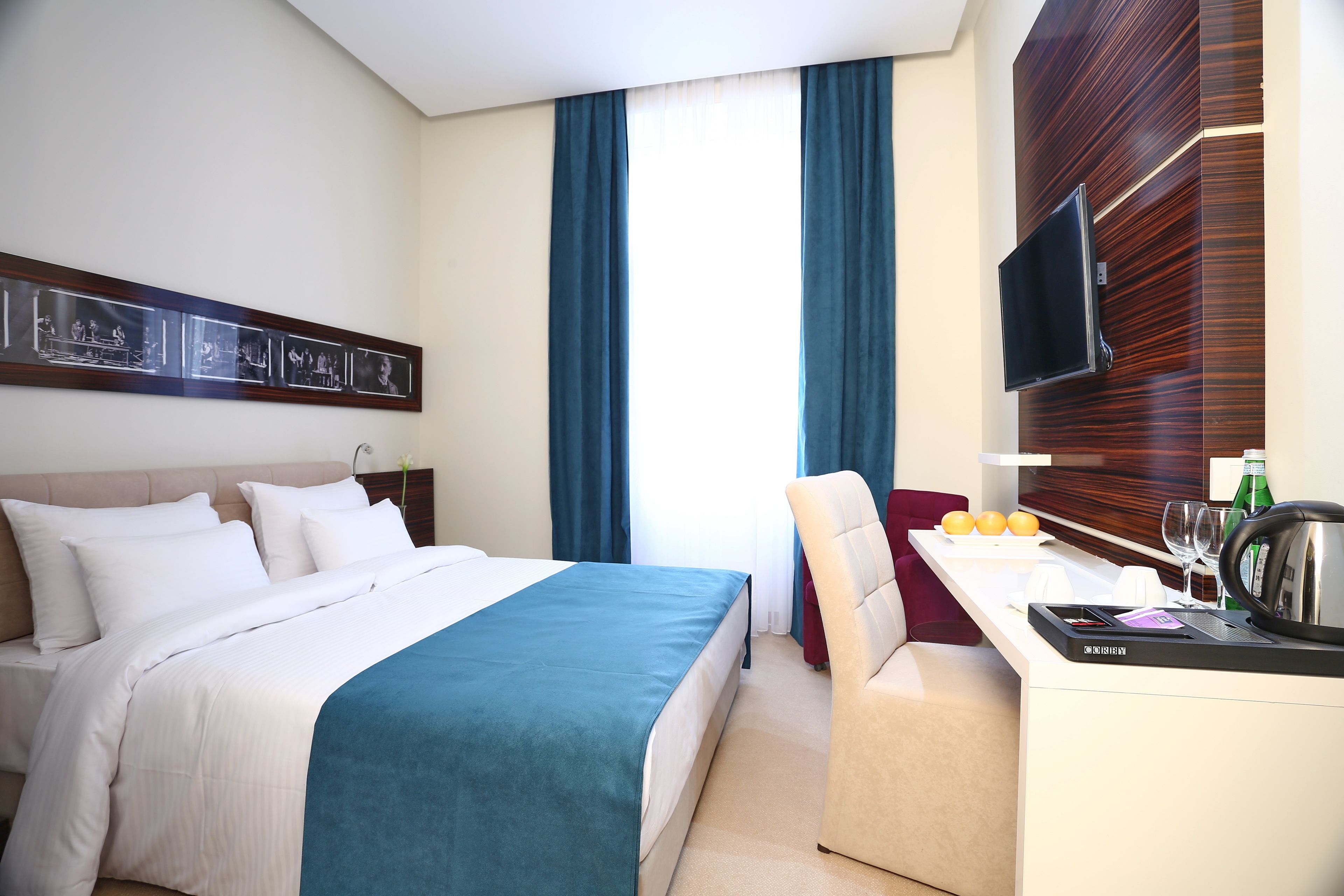 A hotel room with a bed, a desk with a kettle, a chair, a flat-screen TV, and a window with curtains.