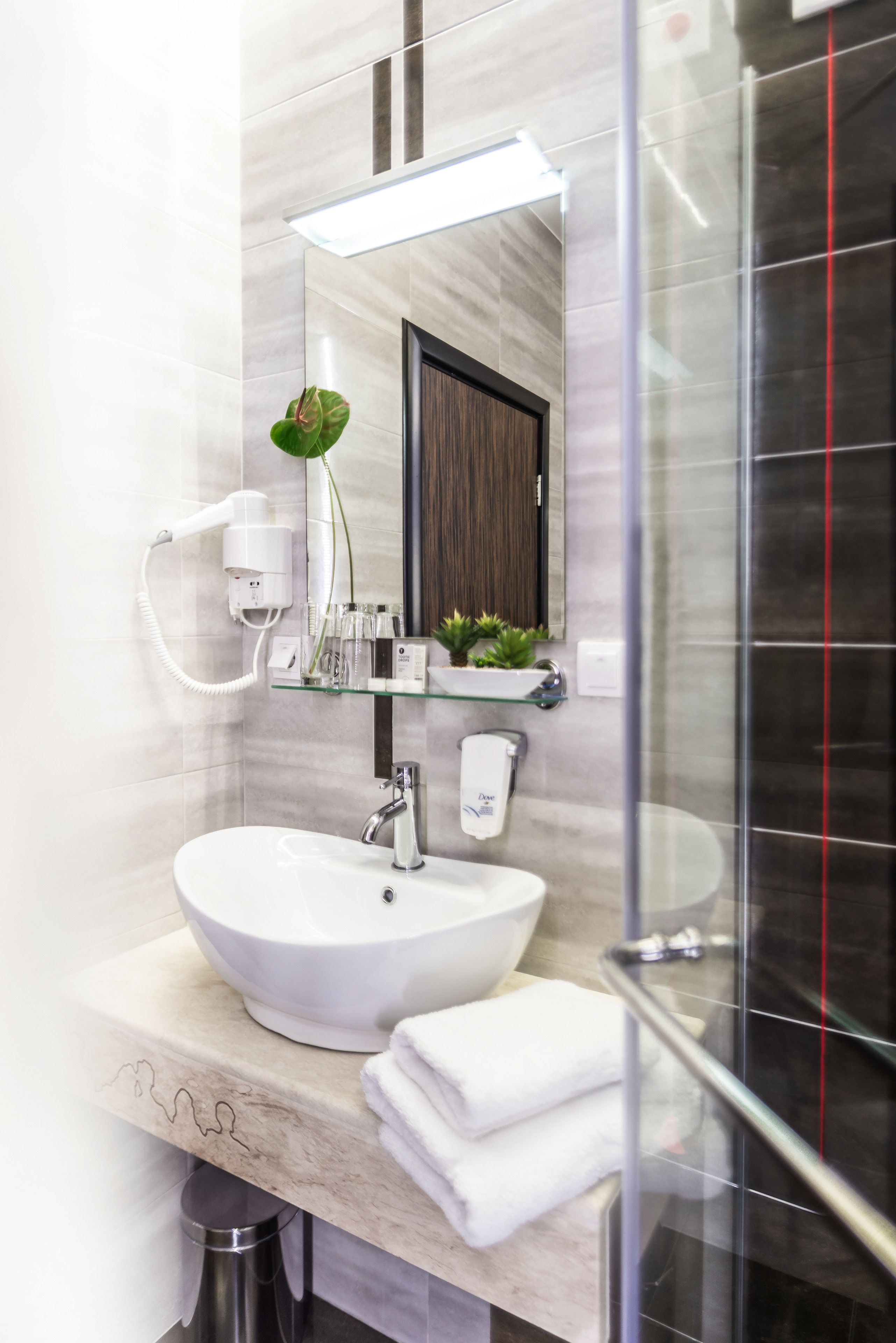 Een moderne badkamer met een witte wastafel, een glazen plankje met toiletartikelen, een haardroger en een spiegel.