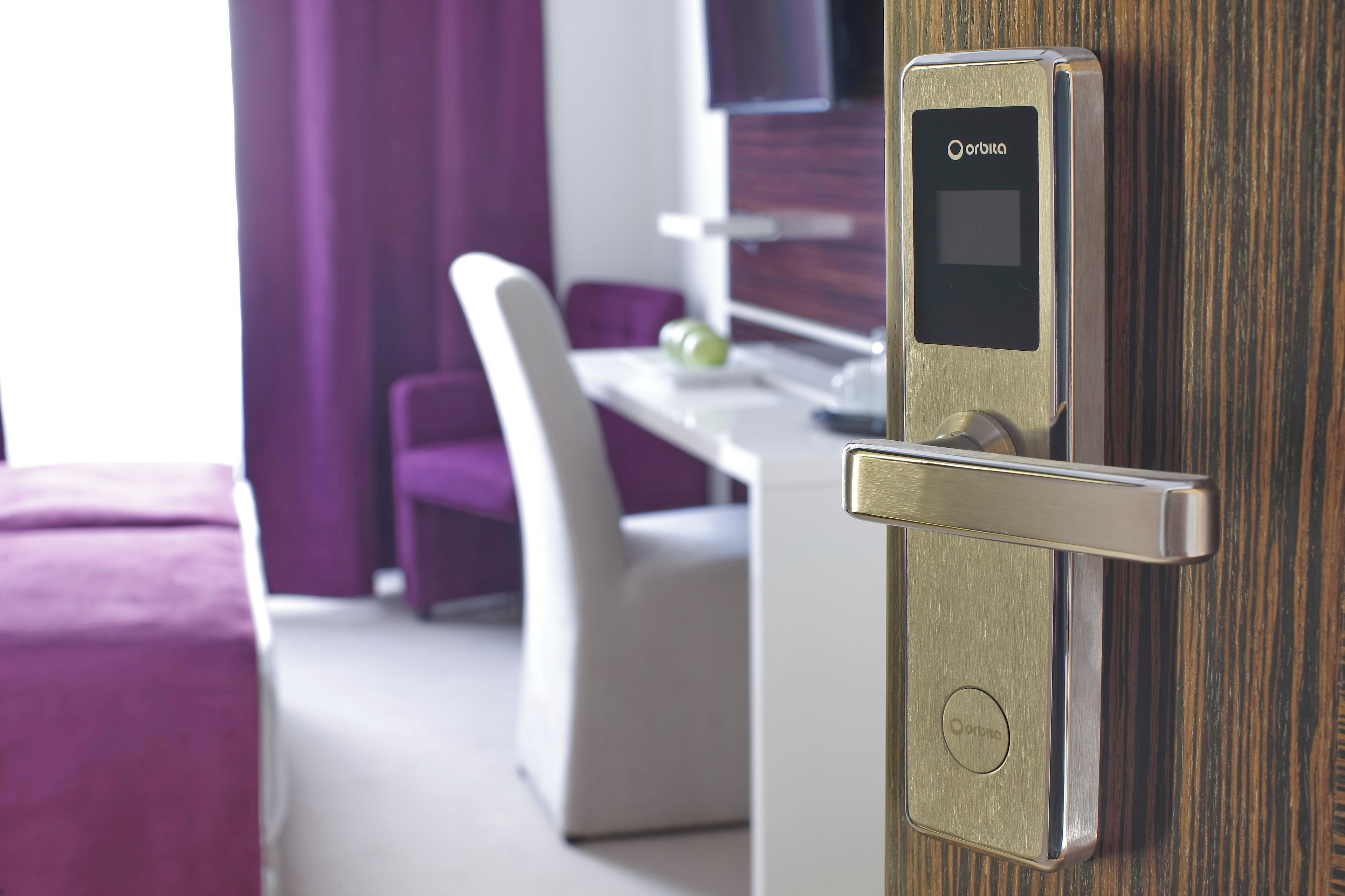 A modern hotel room with a digital lock on the door, a white desk, a purple chair, and a bed with a purple headboard.