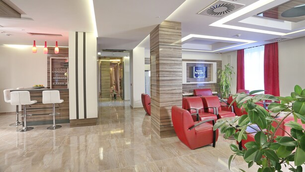 Un moderno vestíbulo de hotel con mostrador de recepción, zona de estar con asientos rojos y una pantalla de televisión.