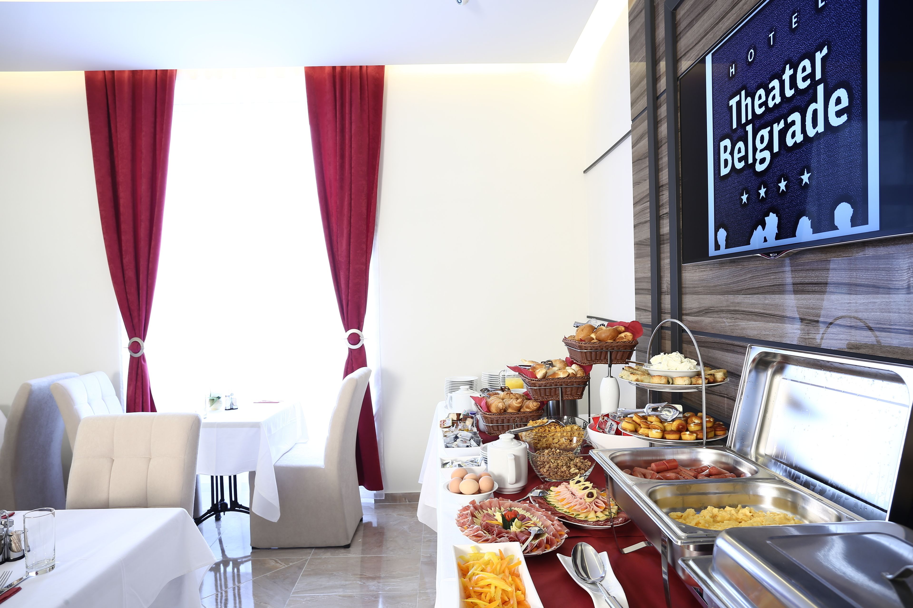 A hotel room with a buffet breakfast setup, a television displaying 'Theater Belgrade', and red curtains.