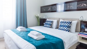 Una cama bien tendida con ropa de cama blanca y azul, una manta turquesa y dos toallas dobladas. Hay una mesita de noche con un libro y un vaso.