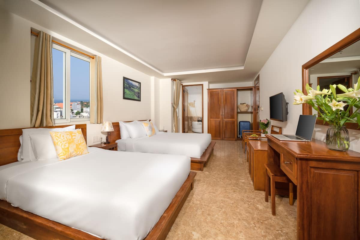 A hotel room with two beds, a desk, a chair, a TV, and a large window with curtains.