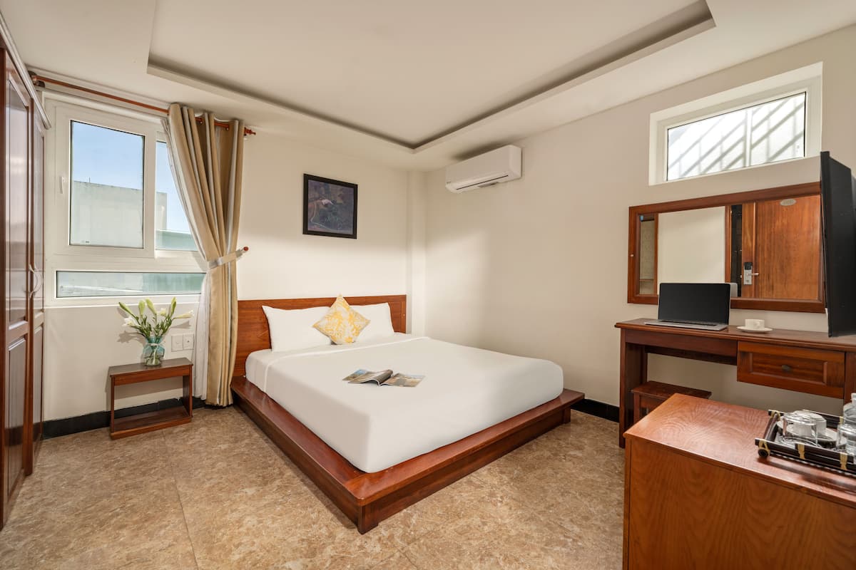 A hotel room with a bed, a desk, a mirror, a window with curtains, an air conditioner, and a small table with a vase of flowers.