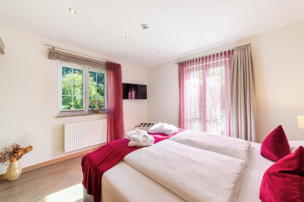 A hotel room with a bed, a window with curtains, a radiator, and a vase with dried flowers.