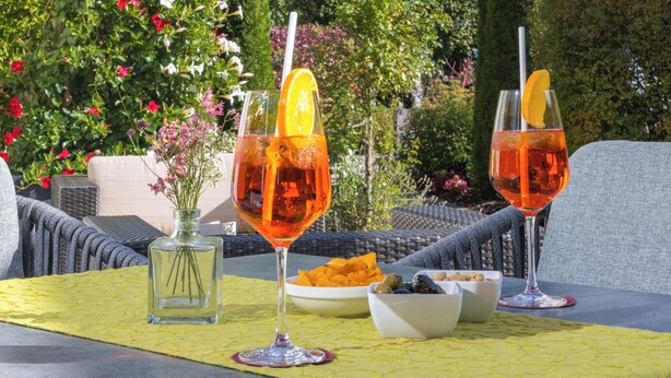 Una mesa con dos copas de vino naranja, un cuenco de frutos secos y un jarrón de flores. Al fondo, un castillo en una colina.