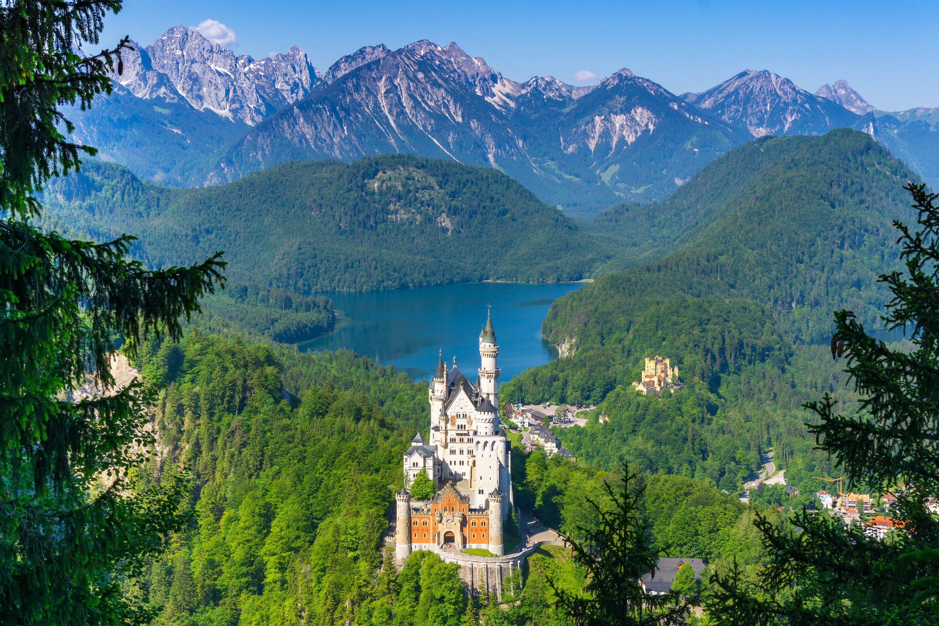 Eine Burg, umgeben von üppigen grünen Wäldern und einem See, mit Bergen im Hintergrund.