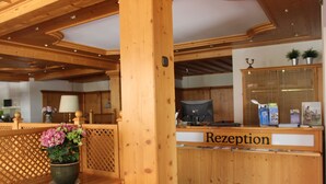 A reception area with a wooden reception desk labeled "Rezeption," a potted plant, and a computer monitor. - Alpenhotel Widderstein (Mittelberg)