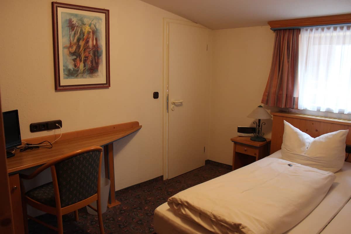 A hotel room with a bed, a desk with a chair, a painting on the wall, a nightstand with a lamp, and a window with curtains.