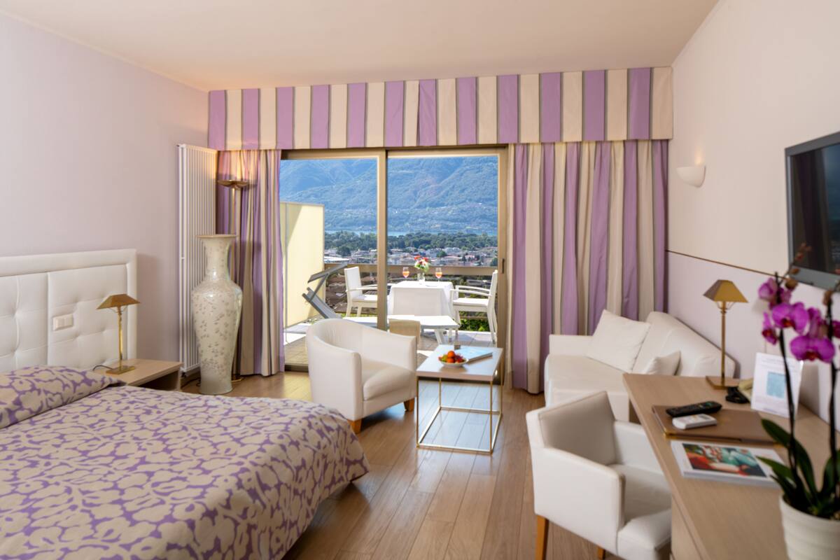Habitación de hotel con cama, dos sillones blancos, una mesita, televisor y vistas a montañas a través de la ventana.