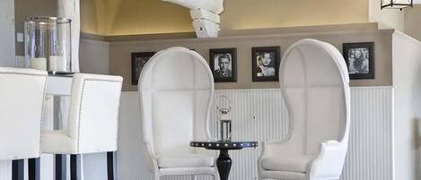 Una habitación con sillas blancas tapizadas, una pequeña mesa redonda con base decorativa y una estructura de columna blanca.