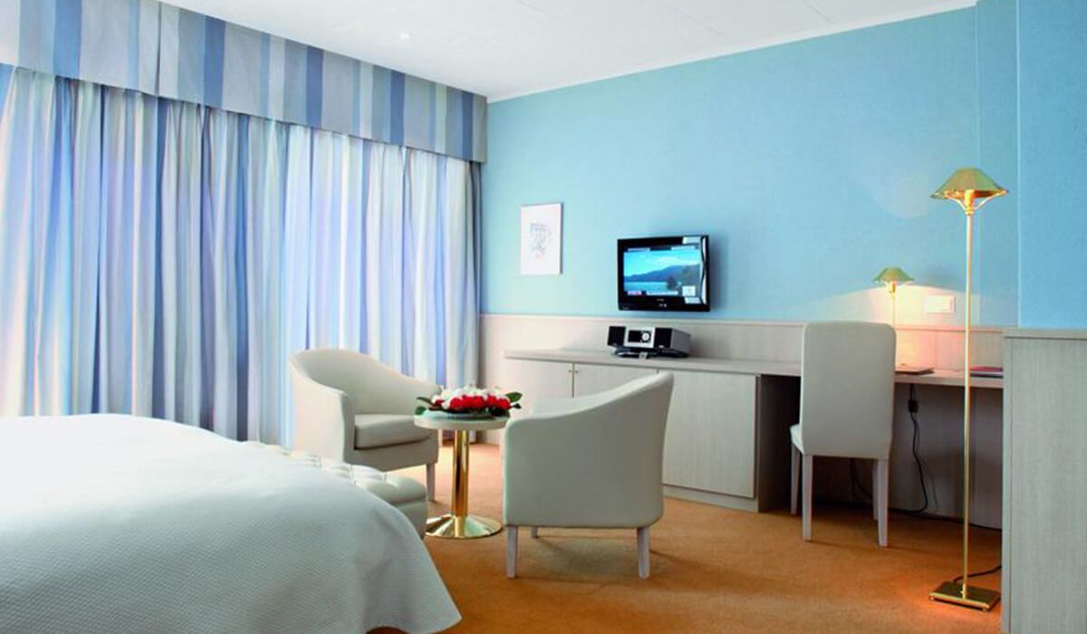 A hotel room with a bed, two chairs, a desk with a computer, a TV, and a lamp.