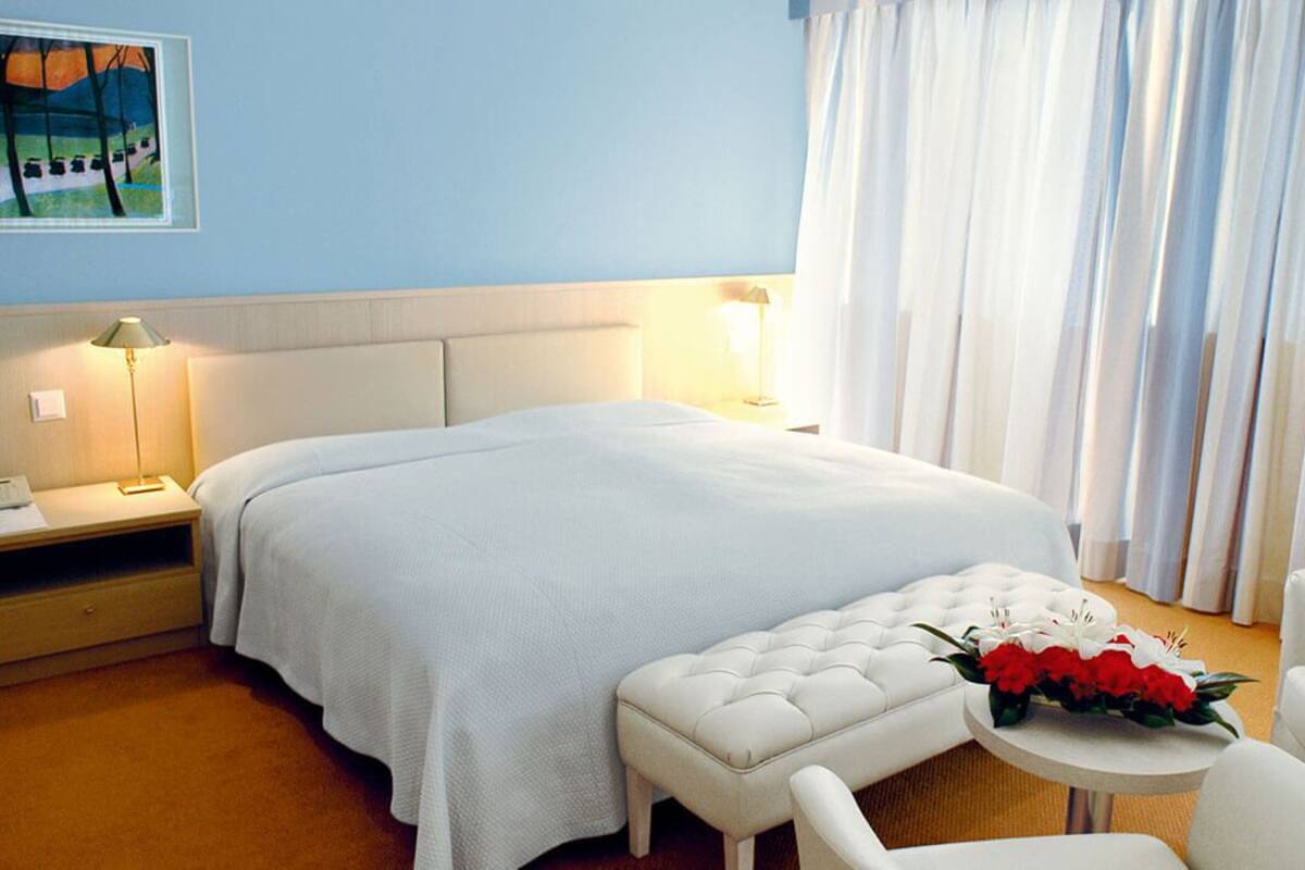 Una habitación de hotel con una cama grande, una mesita de noche con lámpara, una pequeña mesa redonda con un arreglo floral y un sillón blanco.