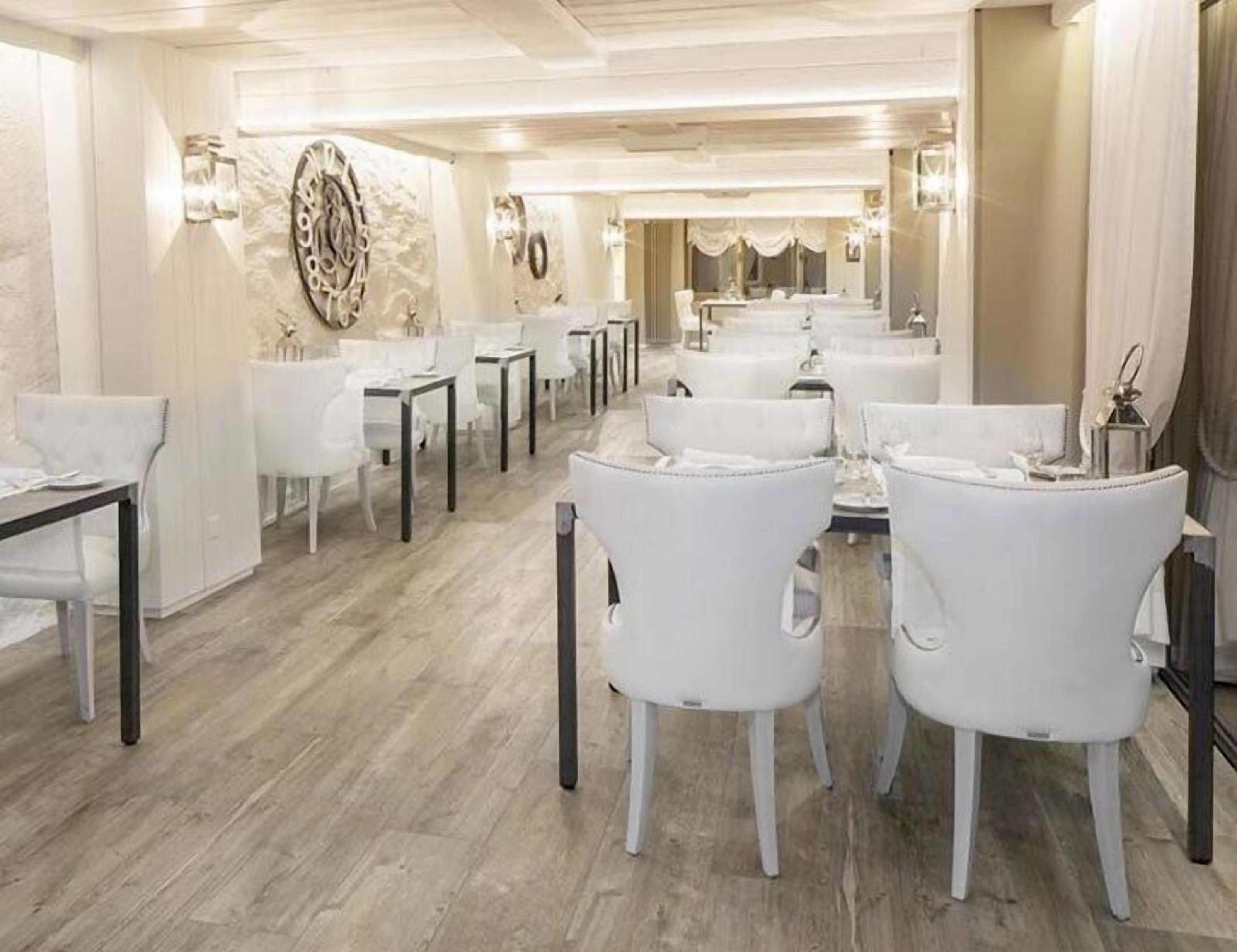 Een restaurantgedeelte in een hotel met witte stoelen en tafels, een marmeren wand met een decoratieve klok en een kroonluchter.