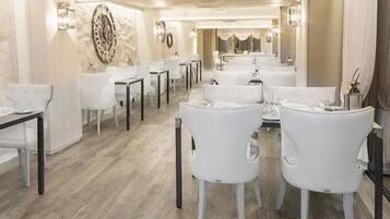 Een restaurantgedeelte in een hotel met witte stoelen en tafels, een marmeren wand met een decoratieve klok en een kroonluchter.