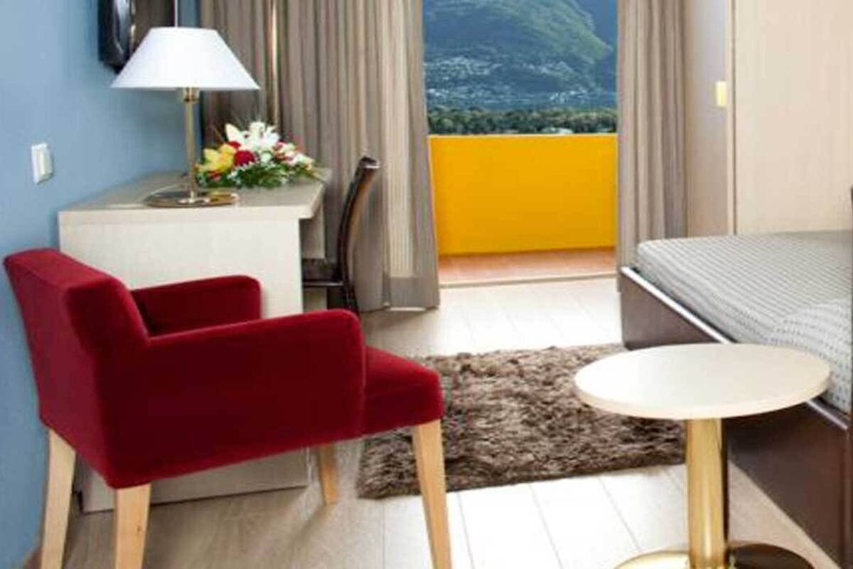 Habitación de hotel con una cama, una silla roja, una mesita redonda pequeña, un escritorio con una lámpara y vistas a la montaña.