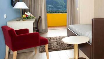 A hotel room with a bed, a red chair, a small round table, a desk with a lamp, and a view of a mountain.