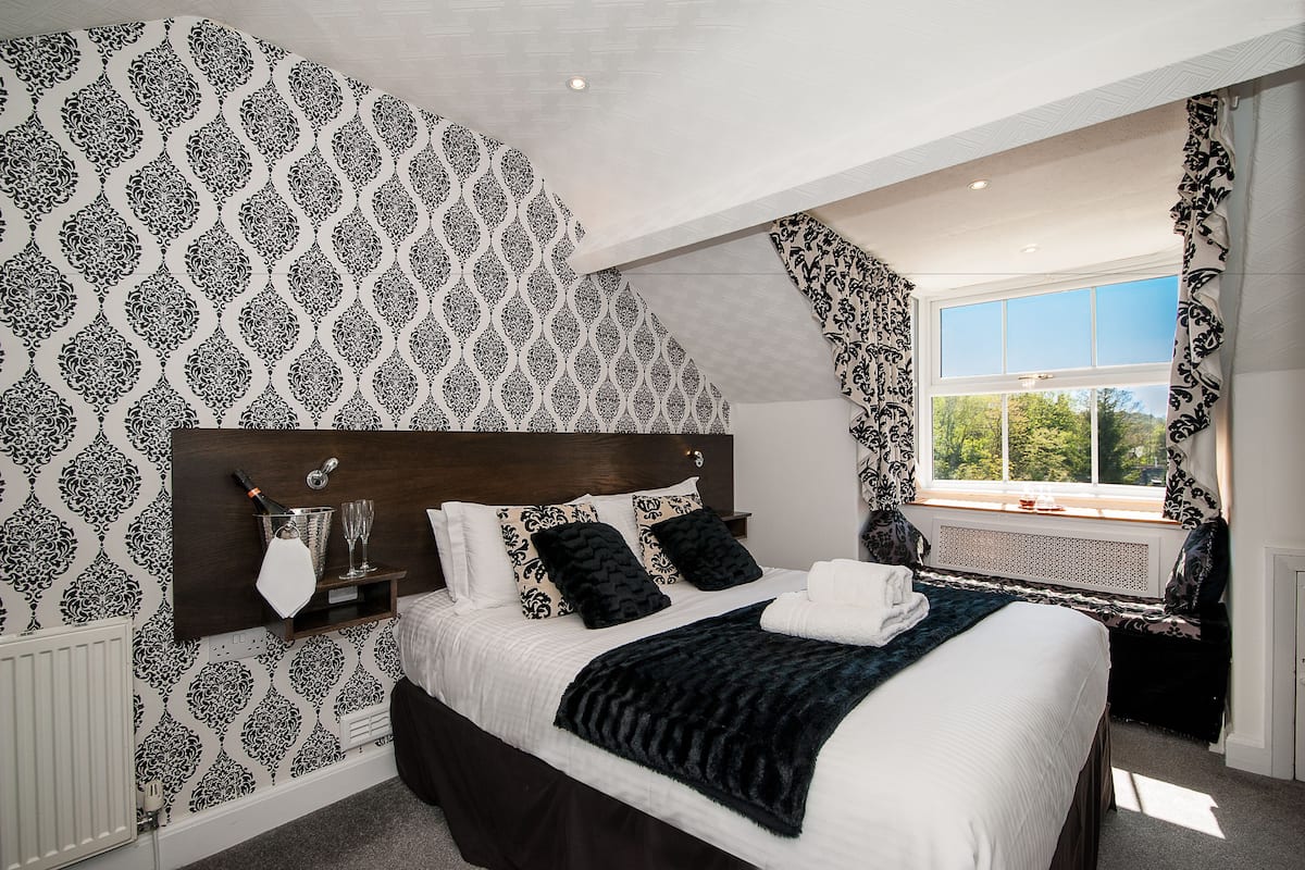 A bedroom with a patterned wallpaper, a large bed with a dark headboard, a window with curtains, and a view of greenery outside.