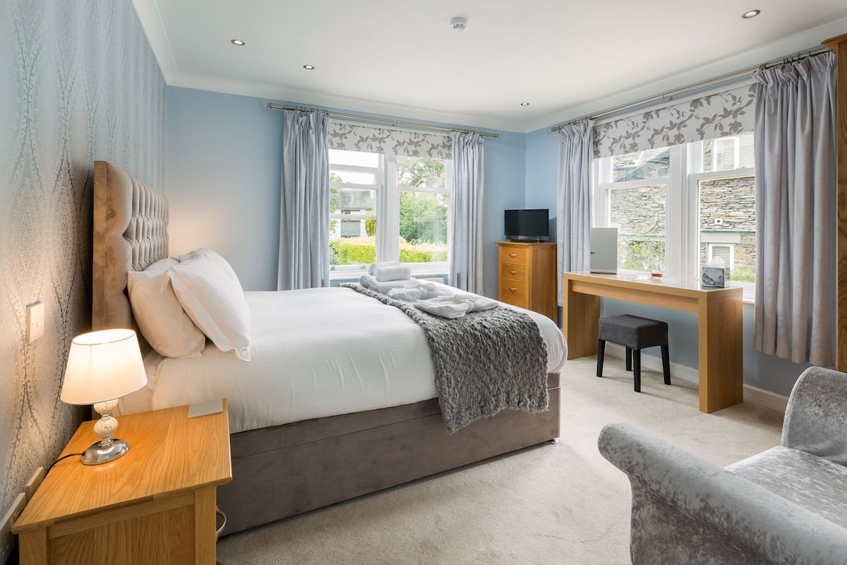 A bedroom with a bed, bedside table, lamp, desk, chair, and a TV.