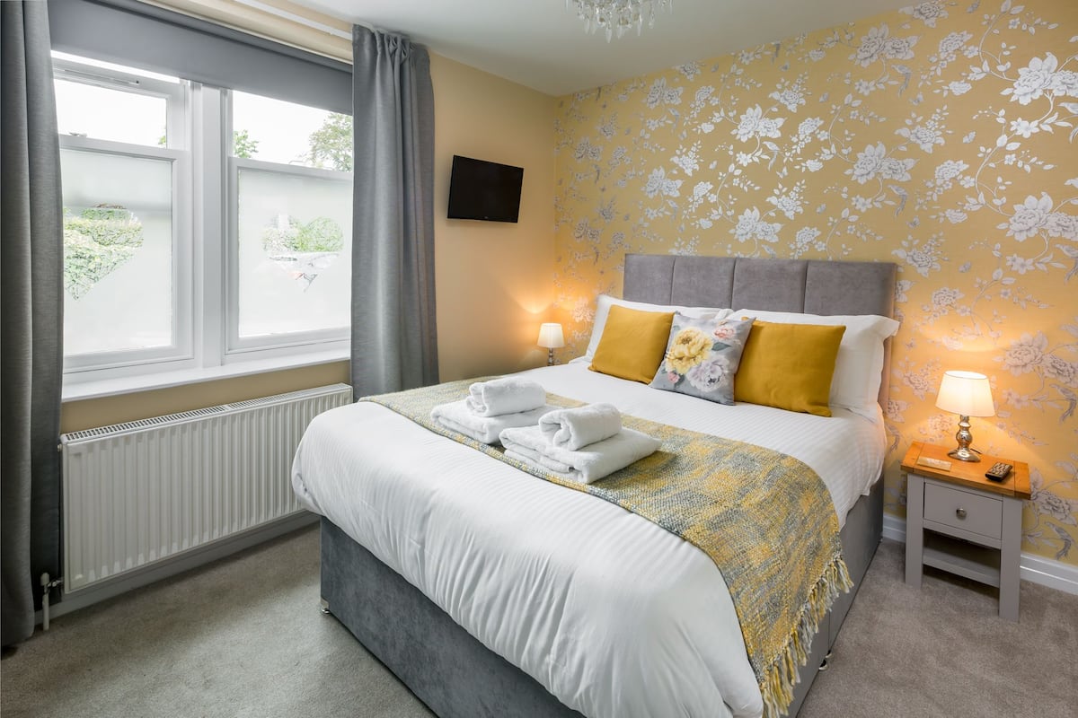 A hotel room with a bed, a bedside table with a lamp, a radiator, and a window with curtains.