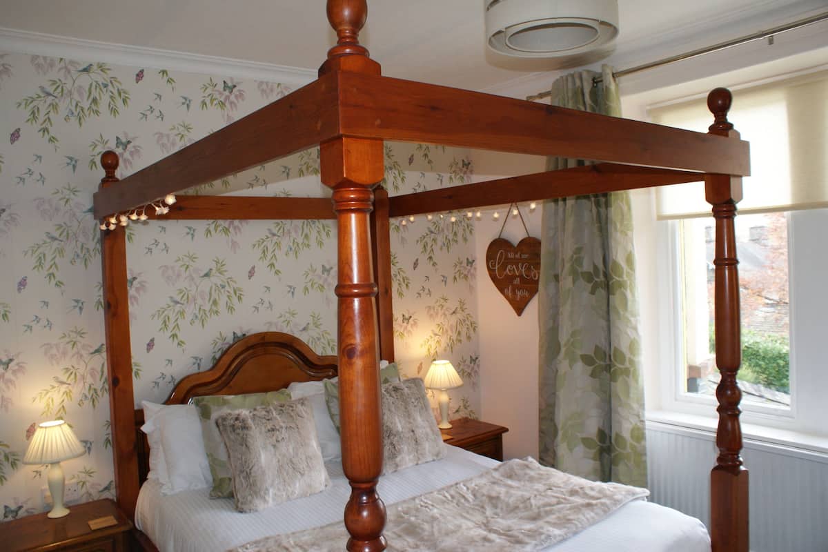 A four-poster bed with a wooden frame, a floral wallpaper background, and a window with curtains.