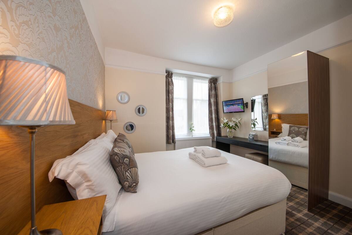 A hotel room with a large bed, a bedside table, a lamp, a TV, and a bathroom mirror.