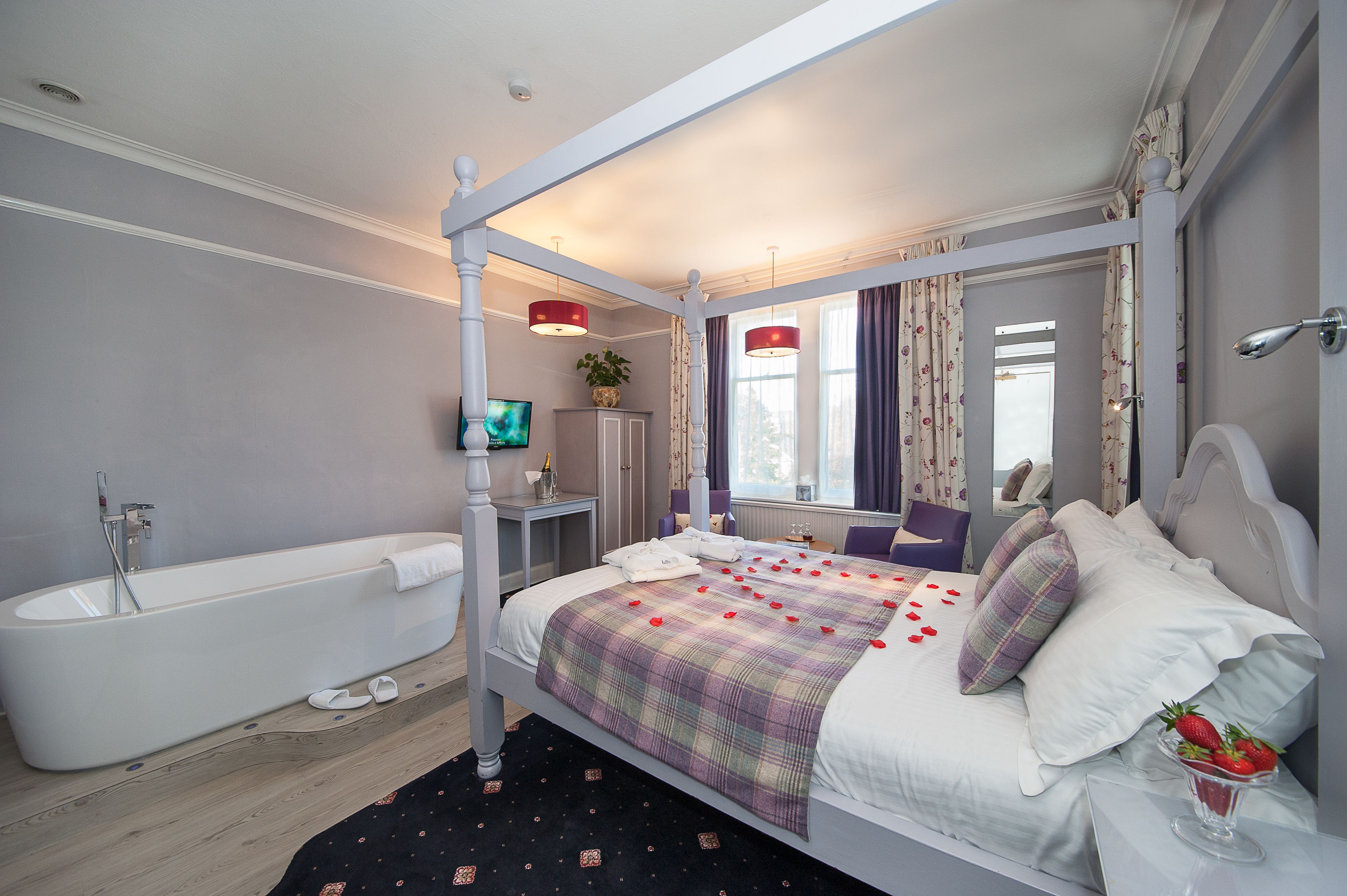 A four-poster bed with a plaid bedspread and rose petals, a white bathtub, and a mirror above the bed.