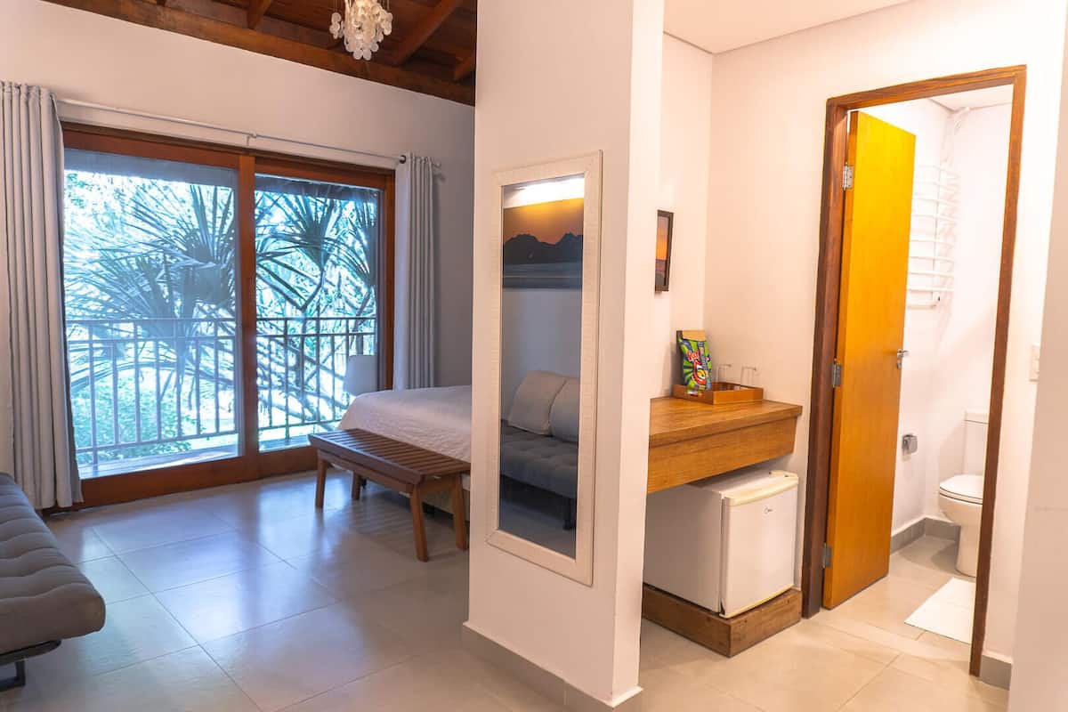Un dormitorio con cama, una banca, un refrigerador pequeño, un mostrador de madera y un baño con inodoro y ducha.