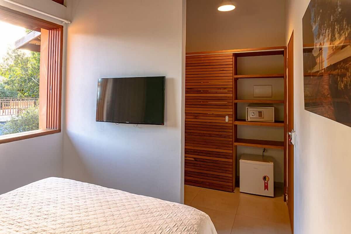 Un dormitorio con cama, televisor montado en la pared, armario de madera y un refrigerador pequeño.