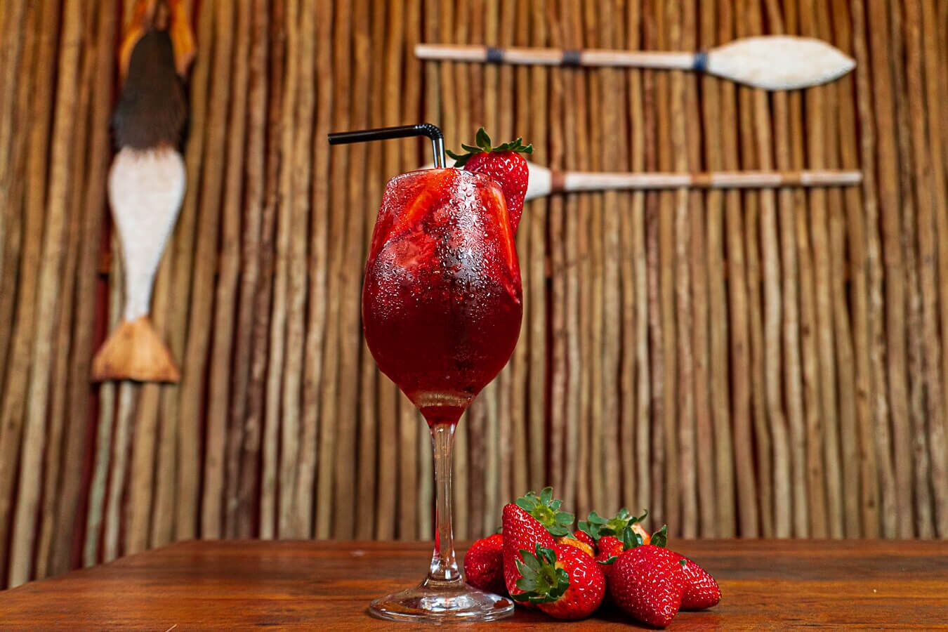 A glass of red drink with a straw and a strawberry garnish, accompanied by fresh strawberries on a wooden table.