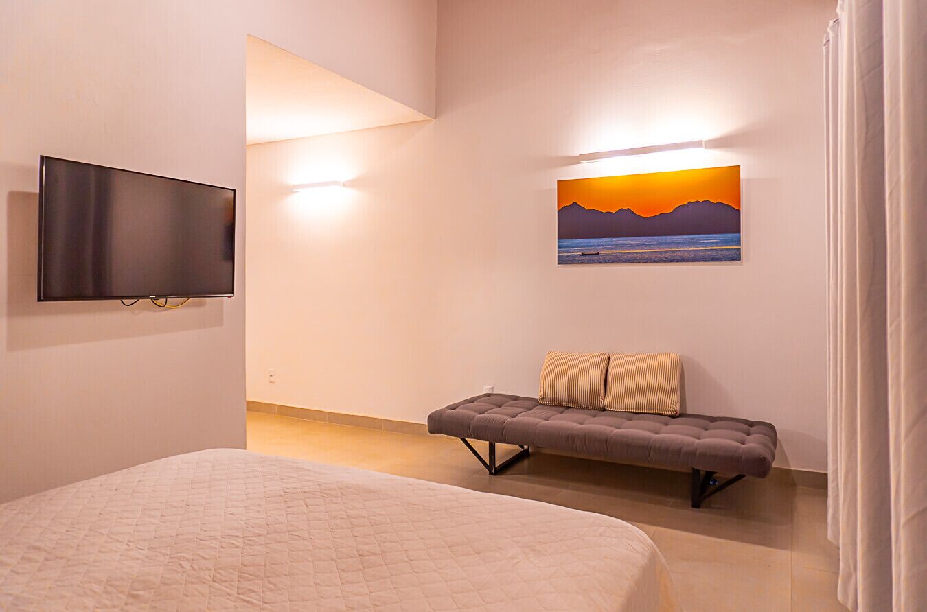 A bedroom with a bed, a bench, a TV, and a sunset painting on the wall.