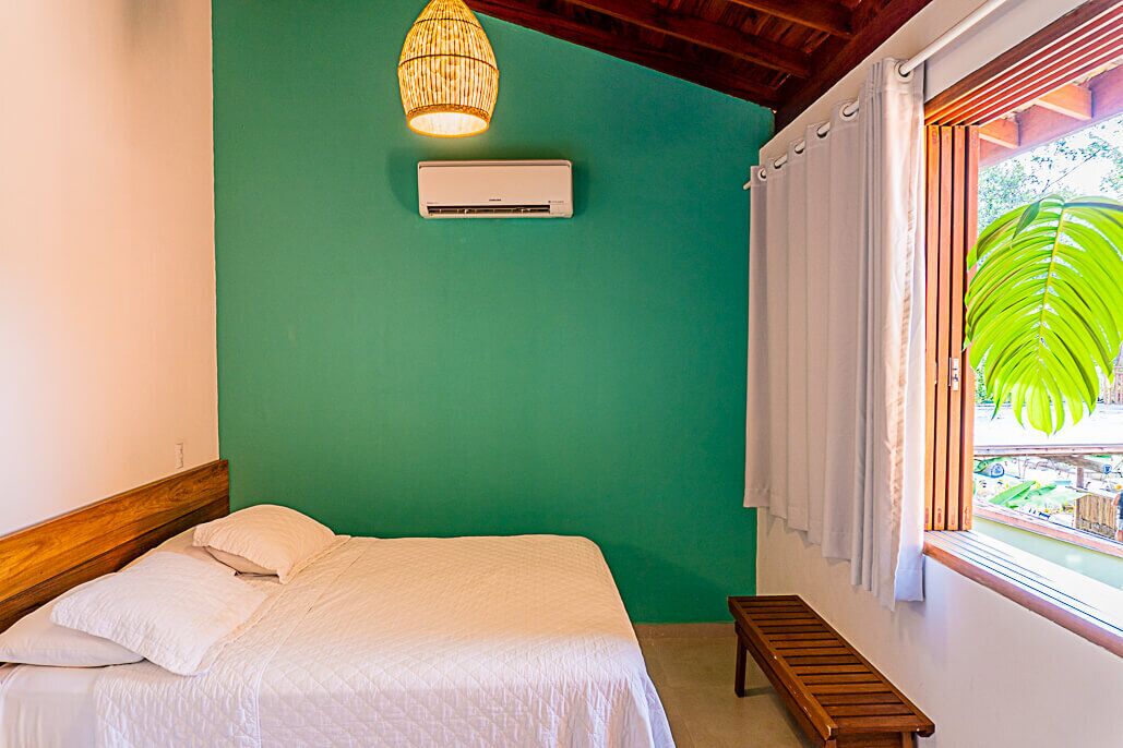 Een slaapkamer met een bed, een airconditioner, een hanglamp, een raam met gordijnen en uitzicht op groen.