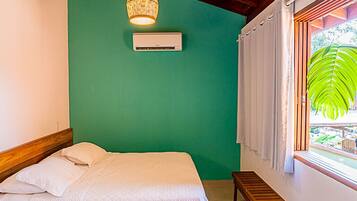 Een slaapkamer met een bed, een airconditioner, een hanglamp, een raam met gordijnen en uitzicht op groen.